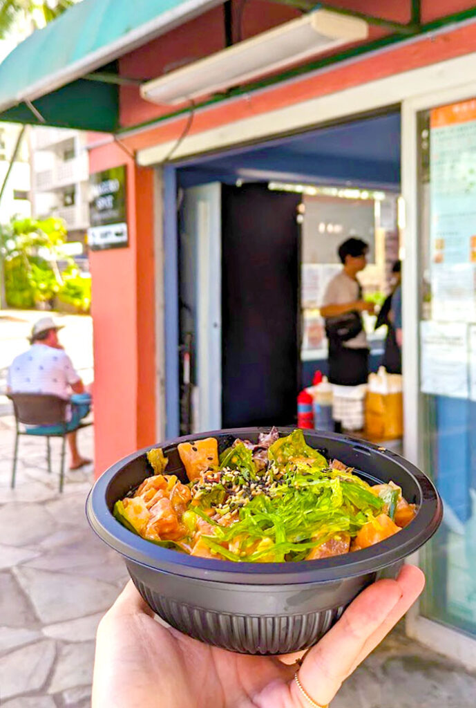 a hand holding up a black bowl filled with poke innside. You can only see the mayo covered salmon pieces, bright green kelp pieces, and furikake sprinkled on top. the entrance to the restaurant is blurred in the background, someone is sitting at a table outside while someone else waits at the cash to pick up their order