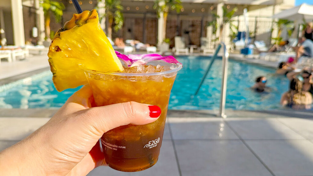 a mai tai on a woman's hand is the main focus of the image. It's a gradient from golden yellow to a darker brown. On the rim of the clear plastic cup is a large slice of pineapple, and a purple lei flower. Blurred in the background is a pool with people in the water. 