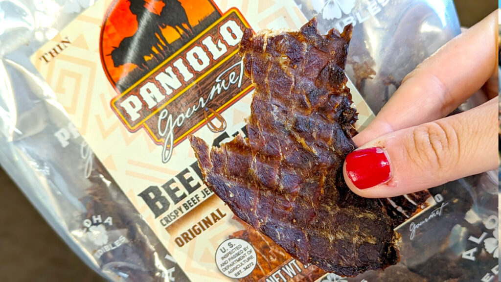 a hand holding a thin beef chip in their hand in front of the packaging that says paniolo, there's a red semi circle with a black silhouette of a horse in front