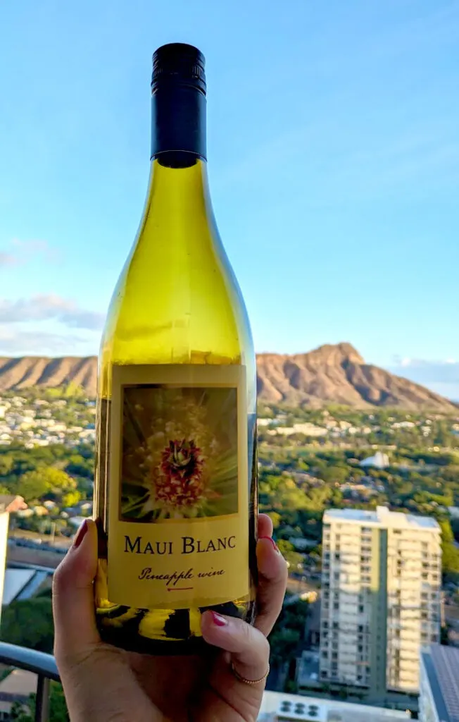 a hand holding up a light green tinted bottle of pineapple wine. In the background is diamond head crater on a sunny blue sky day. The label says: Maui Blanc, pineapple wine