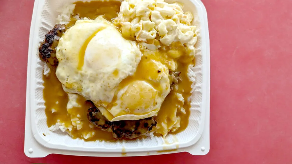 an overhead shot of a traditional loco moco plate lunch in hawaii. A red table provides the perfect contrast background, The food is in a plastic container. The dish kind of looks like mush, but if you look closely, you can see some rice on the bottom, with two grilled beef patties, and two fried eggs. Covering everything is a nice serving of brown gravy, except for the mac salad which sits in the corner on the top right of the dish. 