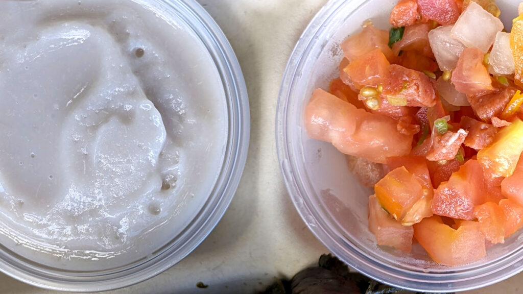 two small clear plastic cups sit side by side in a cardboard container.  the photo is taken from above to see the contents of each cup clearly. The container on the left has a thick purple greyish mash. The cup on the right has a chopped tomato salad, there's supposed to be salmon in there but it blends in with the tomatoes