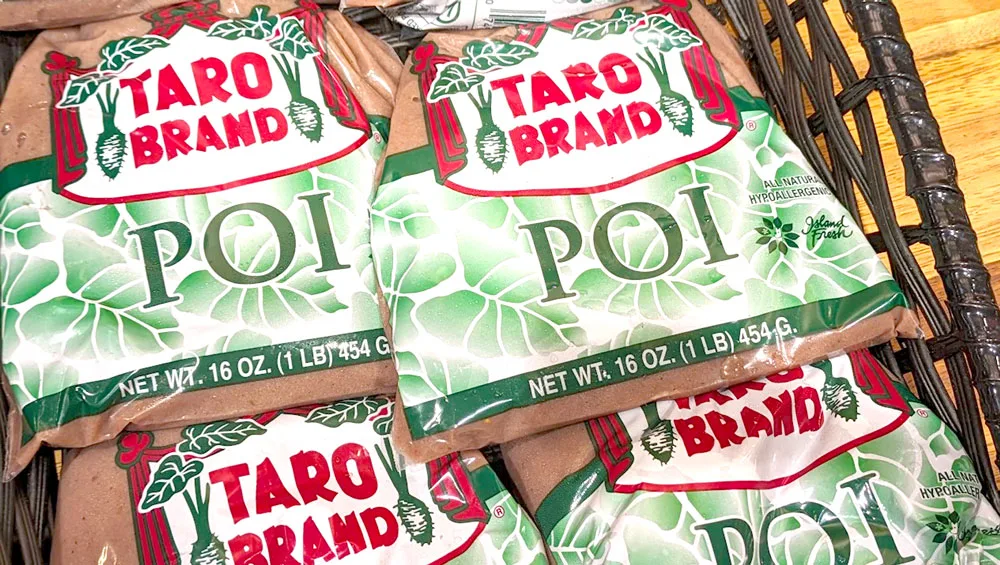 A basket filled with four GIANT bags of Poi. The label says Taro Brand and there are green leaves in the background. The poi is an unsaturated purple and it looks like mashed potatoes.