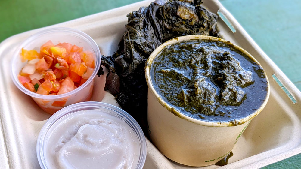 a closeup of a cardboard container with four different types of food in their own container. Starting on the top left, a small bowl holds a chopped red salad. It's a mix of tiny pieces of tomatoes and salted salmon with pieces of onion. To the right is a deep green slimy leaf covering something inside. Below that is a bowl filled a dark green liquid and the same leaf popping out of it. To the left is another bowl container with a purple grey thick substance - poi.