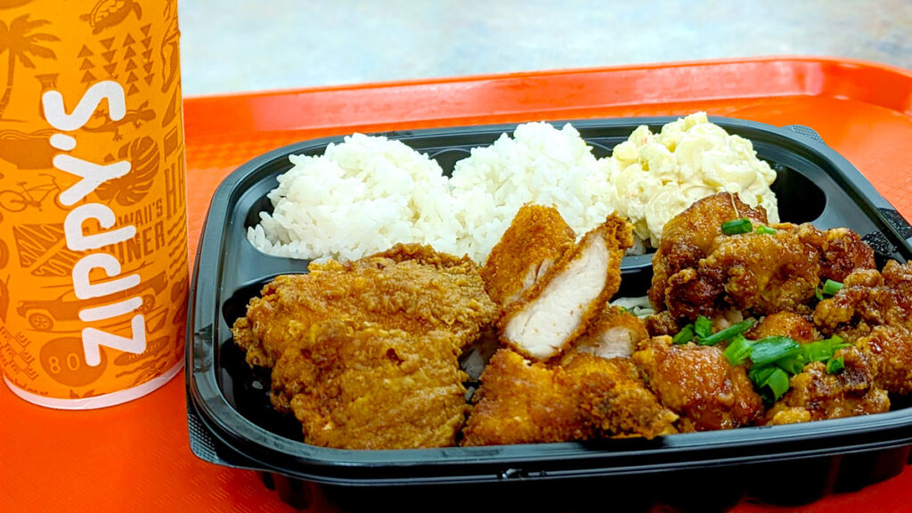 a closeup of the chicken katsu plate lunch from Zippys. There's a soda cup that says zippys on the side in white against an orange background. The food is in a black container on an orange tray. The food is in a bento box style container. There are two bowls of rice, and a third ball of mac salad in the back. The front it taken over by three different types of chicken, chicken katsu, fried chicken, and korean chicken. 