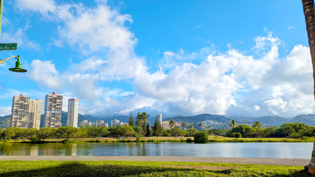 A stark juxtaposition of modern constructions with nature. Grass lines a canal going straight through the image from left to right. Behind the second level of grass in the back is a line of trees, not tall ones, they look more like bushes from this distance. Then there's a layer of buildings, three that even sore high above the mountains that line the background in the distance. The clouds are taking over the peaks of the mountain and leading your eye to the blue sky behind. They also lead your eye to a small sign peaking out from the corner of the left side of the image, attached to an old timey green lampost. The street sign says Ala Wai Blvd