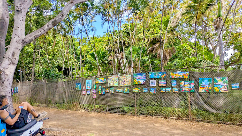 a man in the bottom left corner of the photo looks to be sitting on his motorcycle, but drawing or painting. In front of him is a path before hitting a fence, on the fence there are several art works pinned to it. 