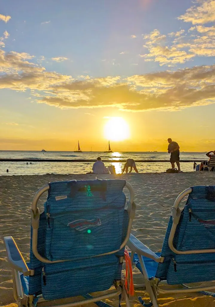 two tommy bahamas beach chairs are photographed from behind. They're empty, but the chairs are on the sand looking out into the ocean with the setting sun. There'a warm yellow glow to the image as the sun meets the horizon .People and sailboats are only a silhouette in the background. 