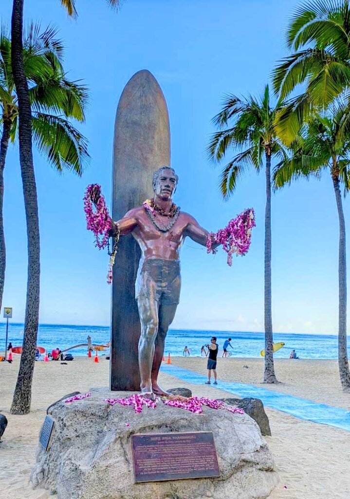 a bronze statue stands in the middle of a large rock  on the beach. The rock is on the sand, palm trees surrounding it, and the ocean in the distance. It's a tall statue of a man in a bathing suit with his arms outstretched. There's an even larger surfboard standing up behind him. Around is neck, on each arm, and on the ground in front of him are purple flower leis that people have left. 