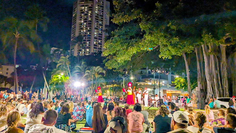 a crowd sits around a stage on the ground. There are two girls in a red dress in front, they are holding hawaiian tambourines. Behind them is a band, dressed in white and playing a ukulele and singing. It's not a completely circular stage, their backdrop is a large banyan tree where the leaves hand above the stage. It's a night time performance