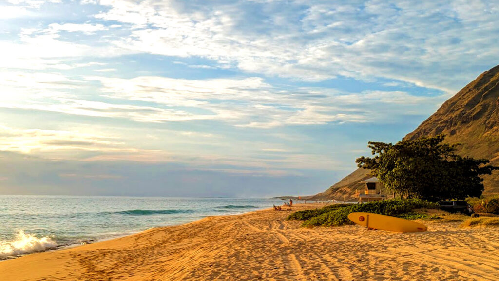 golden hour on oahu at yokohama bay in hawaii. It's a remote part of the island, only a few people are on the beach in the distance. A wave crashes on the baech. There's only a small trip of golden sand on the beach before a rocky dark brown mountain side comes into view. There is still a lifeguard post, almost hidden in the mountainside and behind a single tree on the only patch of grass. There's also a singular surfboard standing on it's side, almost blending into the sand as it has a yellow bottom. It's golden hour, so the sky is a whisp of clouds changing sky colours from a light blue, to a yellow, orange, and pink hues.  