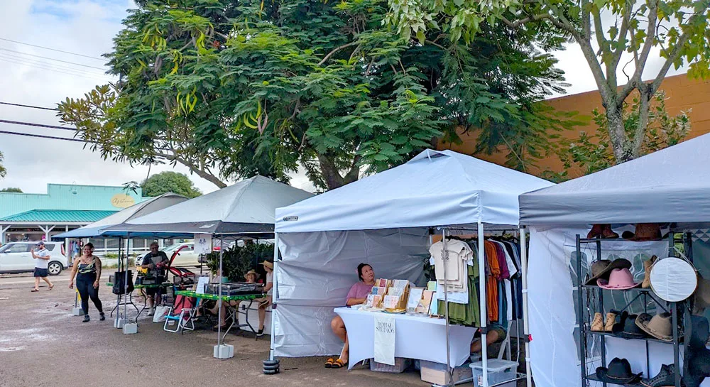 three white pop up tents are set up in a row, each one is filled with different items for sale. The seller sits in their shop as people are walking by.