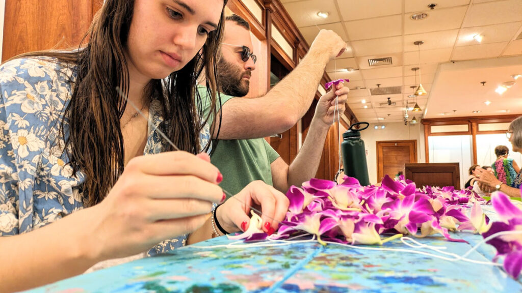 a woman and a man sit at a table side by side, both are in deep concentration. The woman, in the front of the image, is looking down at a pile of purple flowers, she's holding a single one upside down in one hand. In her other hand is a large needle. The man is looking straight ahead as he's holding up a purple flower, pulling a string through the bottom to the top.