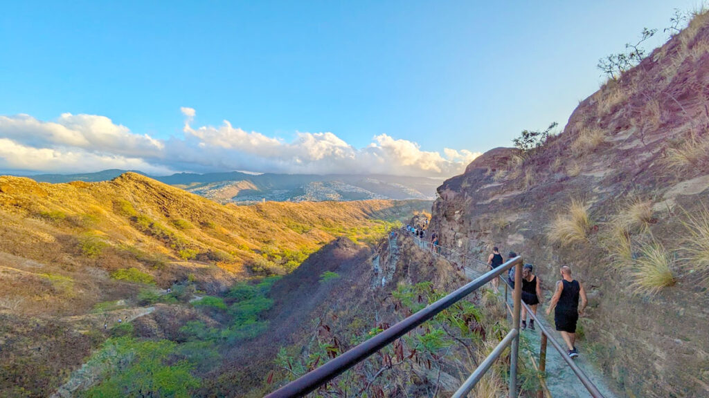 hiking path around the mountain to diamond head. It looks like golden hour with a golden yellow hue to the image, as the sun rises, hitting the mountain side in the distance. The path on the right mountain isn't paved there is a railing, but it's narrow. 