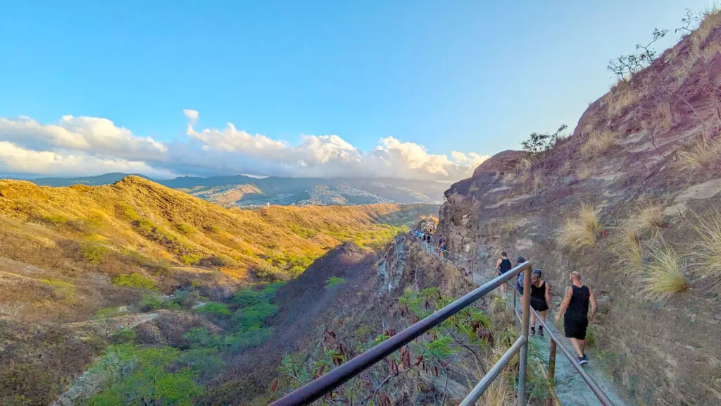 hiking path around the mountain to diamond head. It looks like golden hour with a golden yellow hue to the image, as the sun rises, hitting the mountain side in the distance. The path on the right mountain isn't paved there is a railing, but it's narrow. 