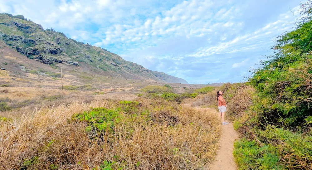 a woman stands further, towards the middle of the photo on a narrow yellow sand path. There is hay like vegetation around her, with some rolling hills and a brown green mountain coming down from the left. The sky is still blue, despite the thick overcast clouds. 