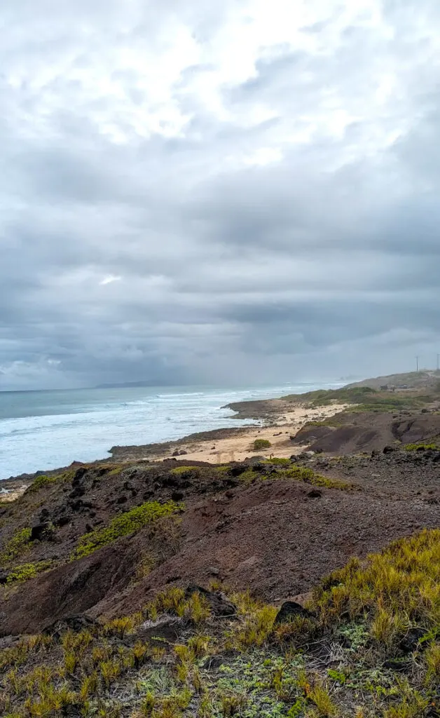 a stark view of the dark, rocky, mars like uneven ground with some specks of green vegetation. This leads into the yellow beach before the blue and white waves crash into the sand. The clouds blend into the blue ocean in the distances. The clouds are thick and great, getting lighter the further away they are from the water. Rays of light try to sneak their way in, but rain in imminent. 