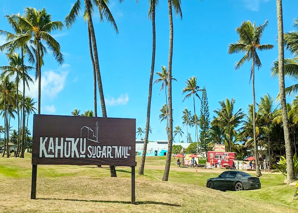 A brown wood sign with white writing stands on the left bottom half of the photo, it reads: Kahuka Sugar Mill. It's on a large fields that's filled with palm trees that are taller than the photo frame. There's one car parks among the rolling hills, and behind that you can see colourful buildings