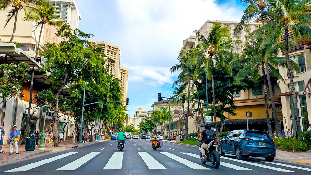 a crosswalk at the foreground crosses a wide street connecting two sidewalks. People are walking on the sidewalks. There are buildings on either side but you can't see what they are because trees are all in front of them. Some buildings do rise above the trees though. On the road, there's only one car, and three motorcycles. One guy on a motorcycle has an attachment to have his surfboard next to him. You know you're in Hawaii when!