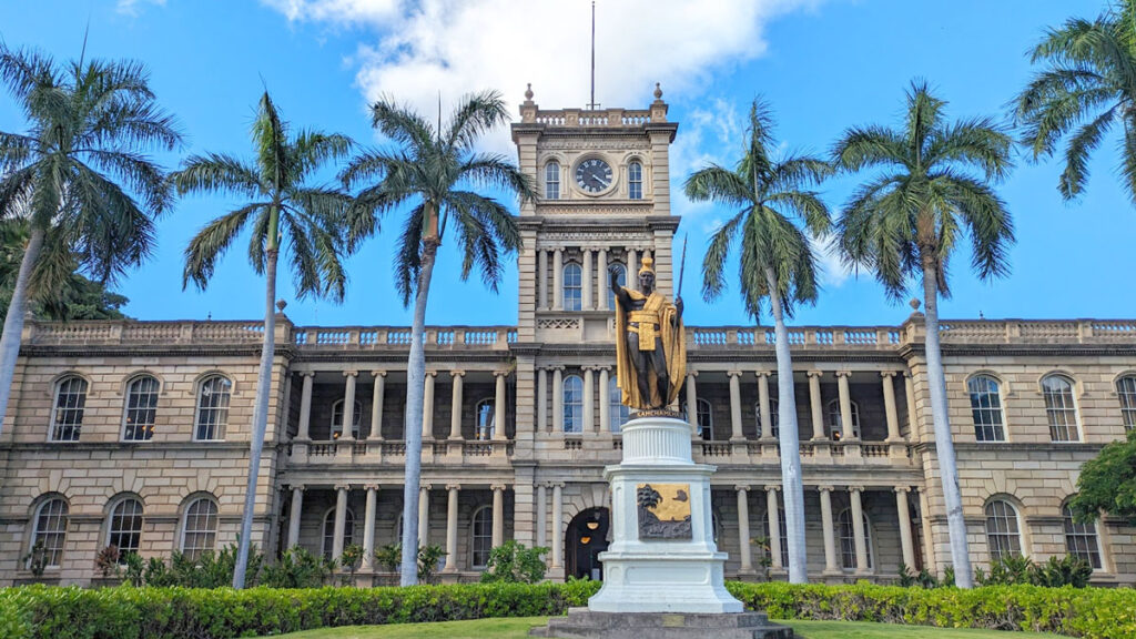 city hall in downtown honolulu a two story building with a centre tower. There are three palm trees on each side of the tower, almost the same height as it. In the centre, on the ground is a gold and black statue of king kamehahmeha in traditional hawaiian clothes. 