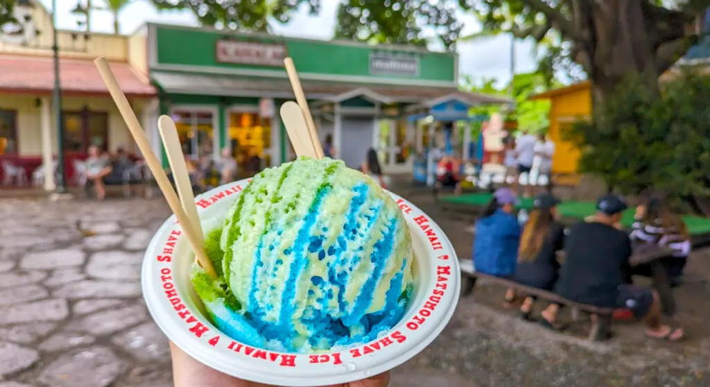 in focuse: a domed blue and green shave ice with a yellowish condensed milk drizzle on top. It's in a paper bowl that says: matsumoto shave ice hawaii three times around the rim in red. There are two paper staws and wooden spoons sticking out behind the dome. In the background is a family of four sitting at a picnic table. There are old timey one story bduilginds painted green, yellow and red. People are blurred walking around, and sitting in front of the shops.  