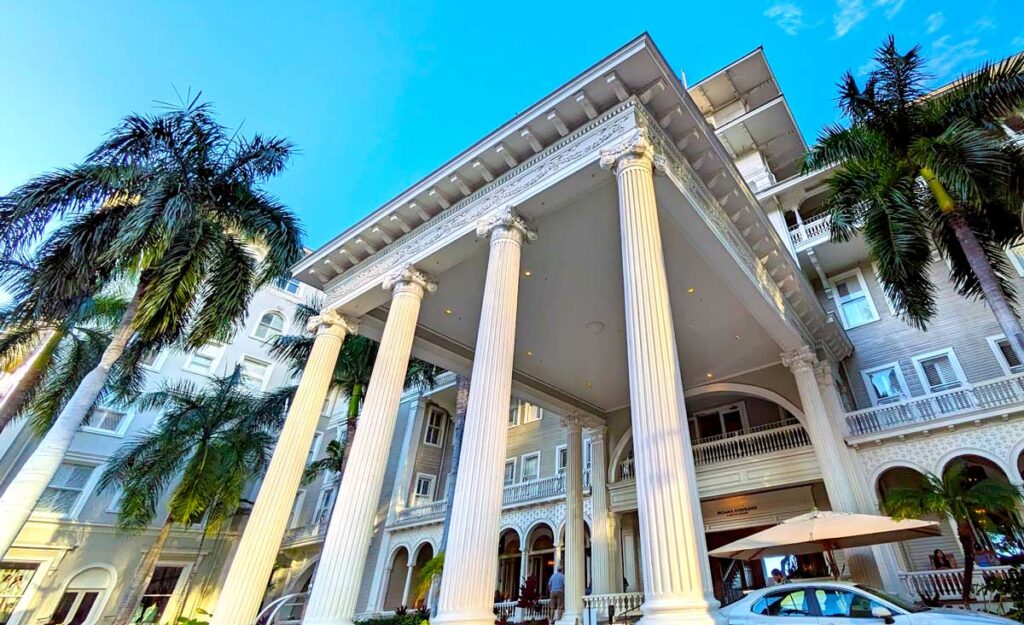 an epic photo showcasing the moana surfrider hotel. The photo is taken from a lower perspective, so the it looks much taller. Four intricate pillars rise up from the ground to support the awning above the wrap around drive way (you can't see it in the photo, but you can see the rooftops of the cars parked htere). The awning leads into the rest of the hotel. It's all in a white stone. There are two palm trees that stand on either side of the building, covering most of the facade. The sky takes up the top left of the image, it a vibrant clear blue. 