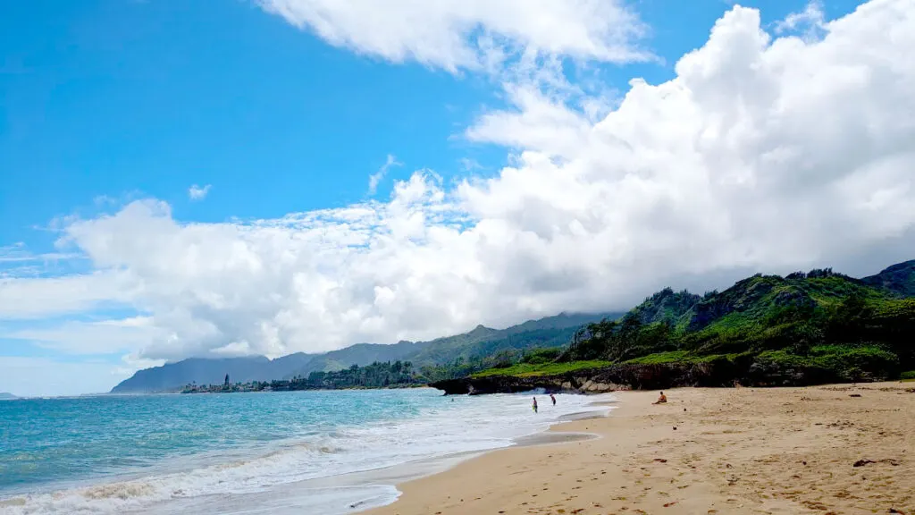 the view at pounders beach. The water is blue, then white when it hits the yellow sand. In the distance, you can see a few specks, these are people on the beach or i the water. The best part about the view are the mountains that line the waterfront and go all the way out. The clouds create an ominous aura to the photo, they are white, but thick coming off the mountains. But there are completely blue skies above the water. 