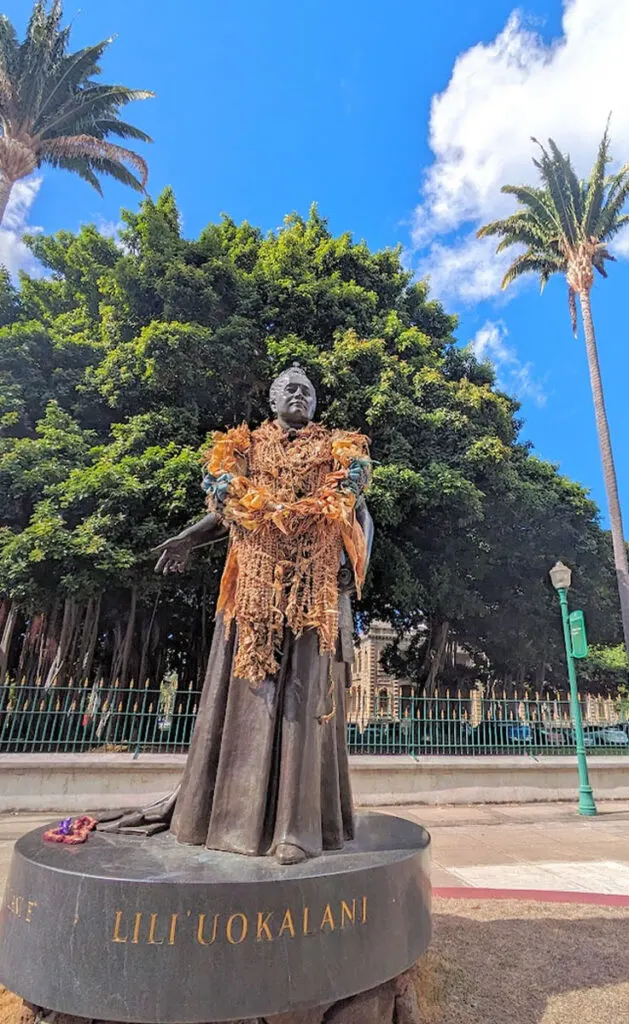 a vertical photo of queen liliuokailani looking powerful and she has old floral leis around her neck, showing how much locals still love here. There's a large banyan tree behind her, and palm trees framing her from above