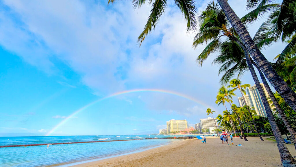 a wide rainbow takes over the sky from behind the buildings all the way to the horizon where the ocean meets the sky. You can also almost make out a second, larger, rainbow above the other one. It's even more faint though. The beach is empty, as it's still early in the morning. The water is calm. Employees are started to set up beach chairs. Palm trees are all along the beach opposite the water before you start to see the buildings and hotels of Waikiki beach in the distance. 