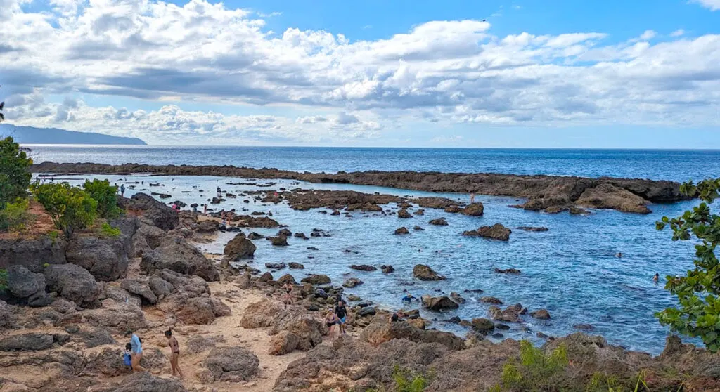 A rocky sharks cove. There's a tide pool that's very rocky with black lava rocks. It's also further separated from the ocean by a nice wall of thicker and bigger lava rocks. People are standing in the water, looking down. The water is choppy though. In the distance, on the horizon is the outline of a grand mountain. 