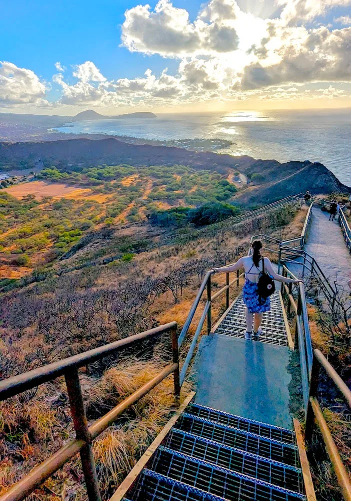 a woman has her back to the camera has she descends metal stairs on the top of the mountain. She's holding on to both railings on either side. She's wearing a blue floral dress, and has a small backpack hanging off of one shoulder. Her hair is up in a pony tail. beyond the mountain and the stairs is teh valley that leads in to the pcean, and the sun is just coming up above the water on the horizon. Casting a wonderful golden yellow hue to the grass and the sky. 