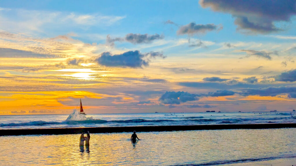 a wave crashes into the waikiki wall at sunset. People in the water are just silhouettes and you can see a sailboat on the other side of the wall. The sky is like a painting, the sun creating a cast of deep orange on the horizon, it gets more yellow before turning blue at the top of the sky. The sky colours are reflecting in the water below. 