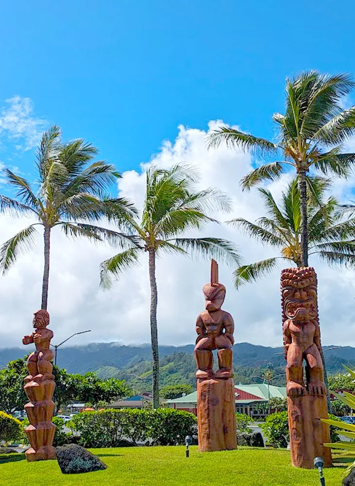 a vertical image showing three tiki statues, all different with scary poses and faces, and each one is a different size. Behind them are taller palm trees, and hawaiian mountains in the distance. There's a layer of thick clouds above the mountain and then completely clear blue skies. 