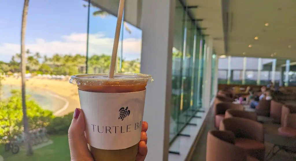 a hand hold outs a milky iced coffee in front of the camera. There's a label on the front that says turtle bay, with a cute turtle logo. Blurred in the background is the cafe, with floor to ceiling windows overlooking the turtle bay public beach. This is one of the best things to do on the north shore