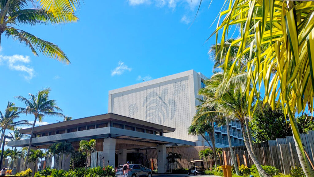 the outside entrance of the turlte bay resort ritz cartlon o'ahu. It has an underpass for people to drive under and be in the shade, or out of the rain. The side of the building is grey concrete, but in a darker grey you see the silhouette of three turtles. The shells of the turtles look like palm tree leaves. Palm trees also surround the building, and frame it.