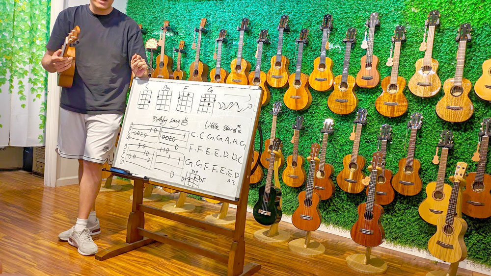 a classroom setting for ukulele lessons. a man holding a ukulele stands next to a white board with guitar tabs and notes, there are a lot of letters in a row from A to G. These are the chords needed to play Happy Birthday and Twinkle Twinkle Little Star. Behind him is a wall filled with all the ukuleles imaginable. 