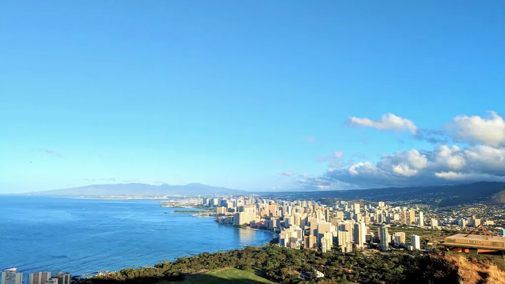 the jaw dropping view from the top of diamond head crater, a city skyline line the beach, where the ocean meets the land. Behind the buildings are greenery and the start of large mountains. There are also mountains forming in the distance. Clouds are coming off the mountains on the right of the image, but otherwise the sky is empty and bright blue.