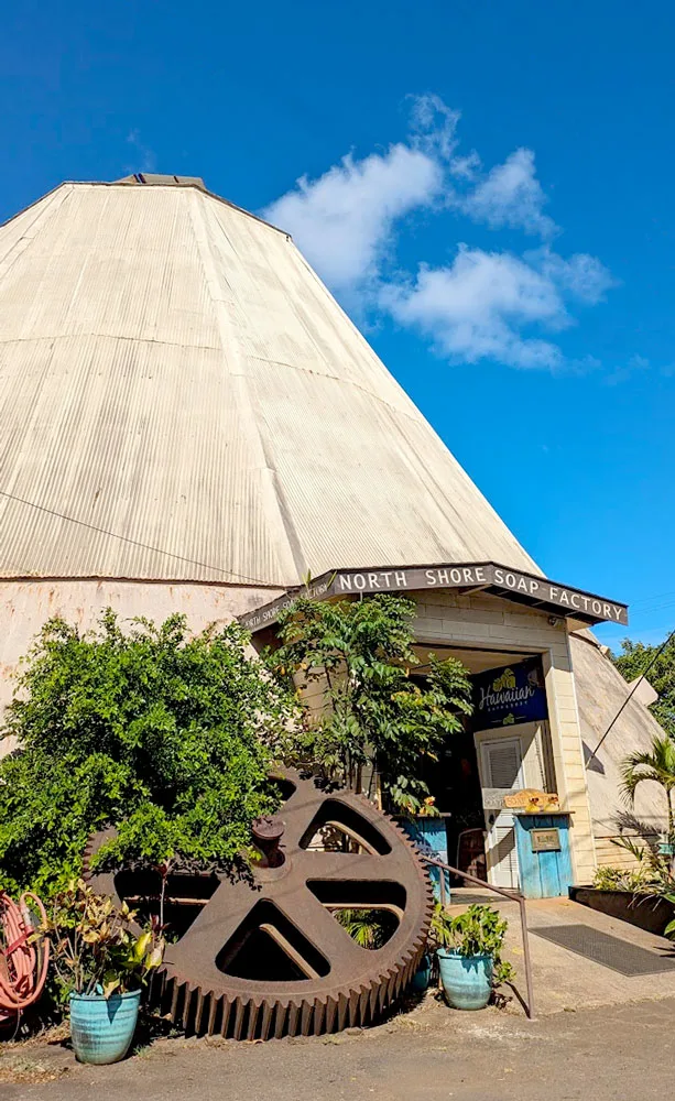 a vertical image of the old waialua sugar mill. it rises high into the sky, on the front it there's a door with the sign for the north shore soap factory. There's an old rustic pinwheel leaning against the mill.