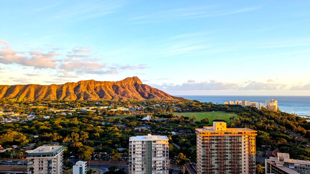 a beautiful view from the 30th floor of diamond head crater during golden hour. You know the view is from high up because you can just see the top of other high rise buildings. There's a valley between the buildings and the jurassic looking mountain that looks golden brown being lit up by the setting sun. 