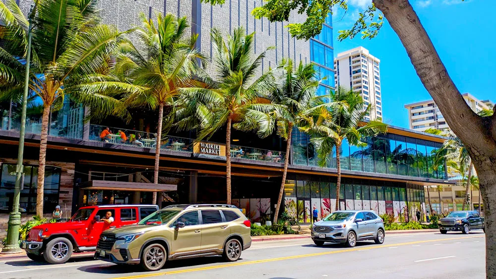 An outdoor view of the waikiki Market on the street. Cars are driving by, there's even a bright red jeep adding a nice pop of colour to the photo. Palm trees stand in front of the waikiki market. It's on the second story of the building, there are blue reflective windows except for in the middle which is all open air. You can make out some tables, and people are sitting at them. 