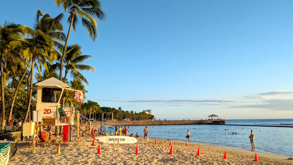 the safety of waikiki beach, a white life guard tower looks out onto the water, there are palm trees behind it , lining the entire beach. Resting below it on the sand is a surfboard that says rescue on it. Cutting the water through the middle is a large stone wall, it also wraps in front of the beach, cutting all the waves that reach the shore in this area.