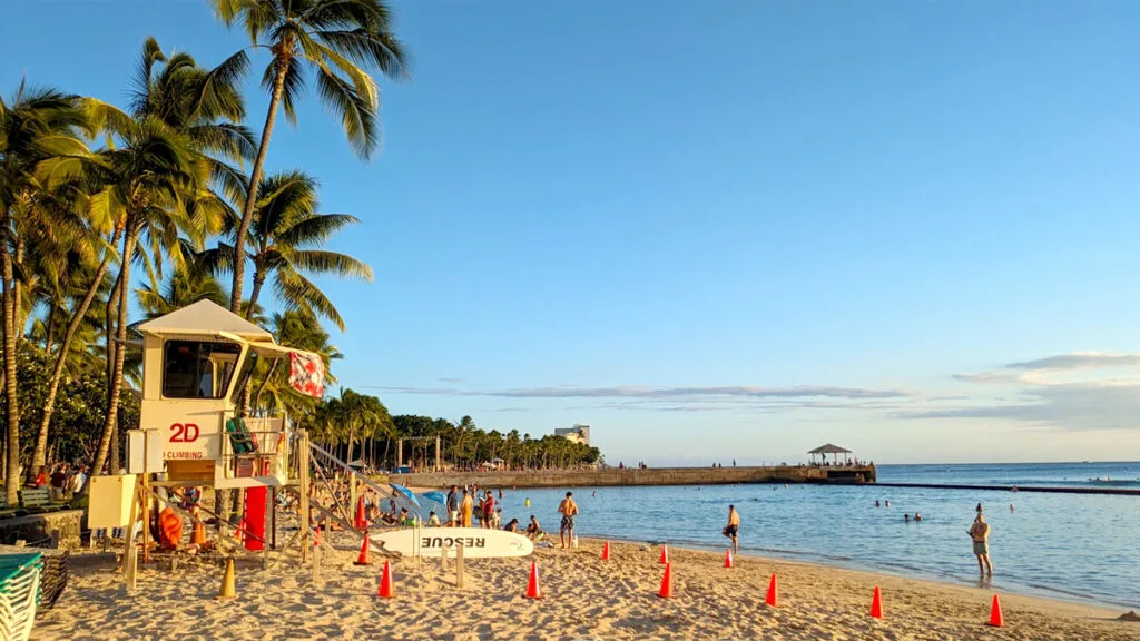 the safety of waikiki beach, a white life guard tower looks out onto the water, there are palm trees behind it , lining the entire beach. Resting below it on the sand is a surfboard that says rescue on it. Cutting the water through the middle is a large stone wall, it also wraps in front of the beach, cutting all the waves that reach the shore in this area.