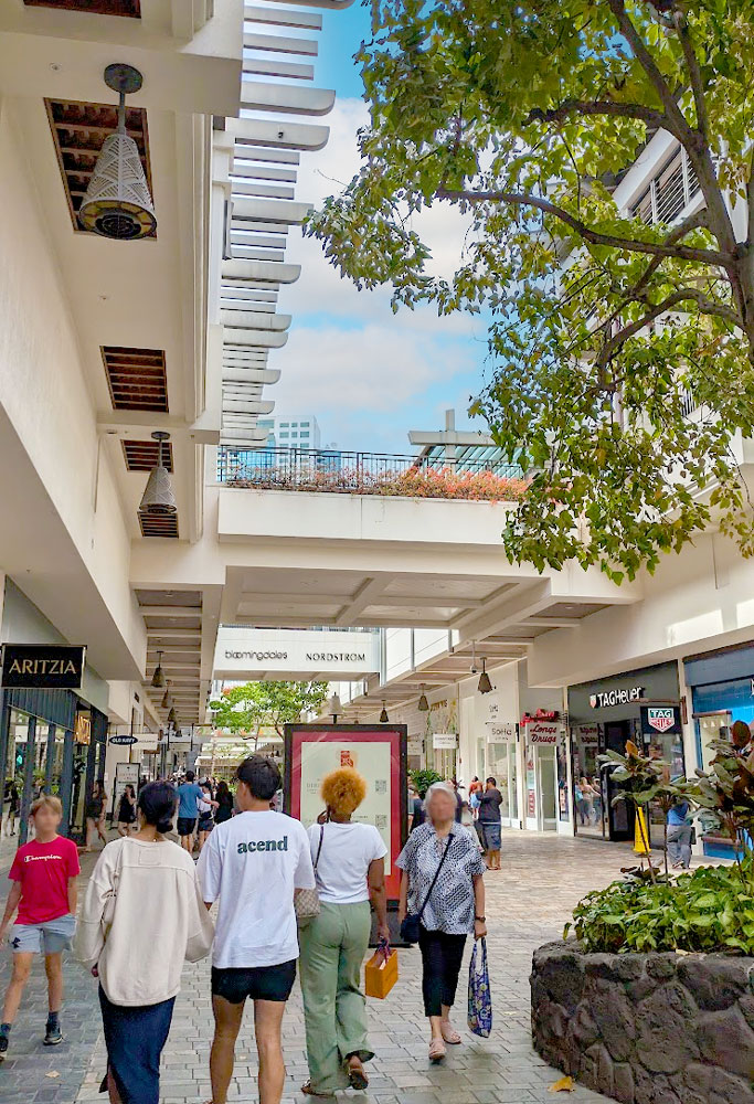 a vertical image of inside a shopping mall hallway that isn't inside at all. You can see the storefronts on either side of the hall, people are walking in the middle. There are plants all around. And you can also see the blue skies above. There's a walkway on the second level adding a bit of shade, connecting both side on the second level. 