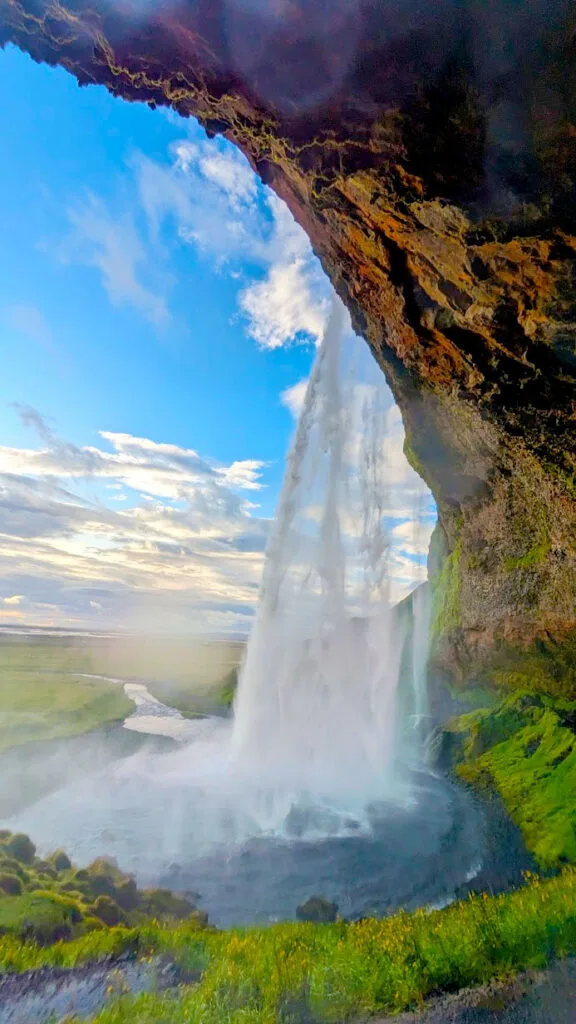 A waterfall reached farther down Icelands ring road a jaw dropping view of a a path that goes in and out of the frame. There's a rock mountainside above where a waterfall is coming down off of it going into a pool of water below that leaves through a narrow stream into the distance. It's the start of sunset, with blue skies, but a golden yellow hue on the horizon. 