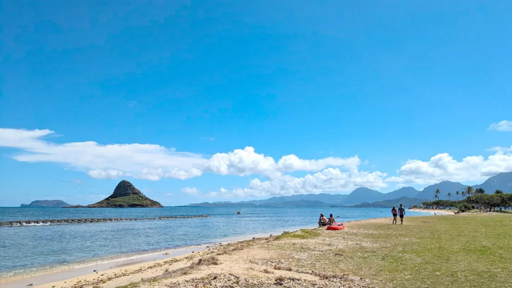 a relaxing hawaiian beach with mokolii island, or china mans hat in floating out in the water. There are some people on the island, but it is far from busy. Behind the island you can see the silhouette of the Windward Coast mountain ranges. Clouds lead off the land and separate and become more sparse over the ocean, leaving bluer skies