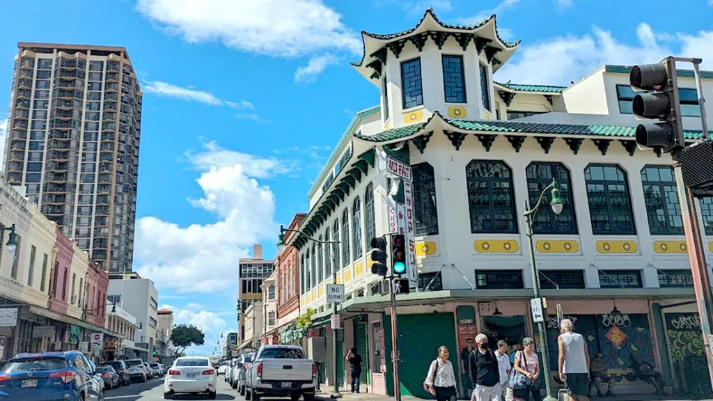 a street in chinatown honolulu. A large three story building is on the corner of the street, it has a circular tower no one side. It's painted white, with a green roof, and gold decals. The roof is a curved pattern. Across the street is more typical buildings that you would see in downtown honolulu