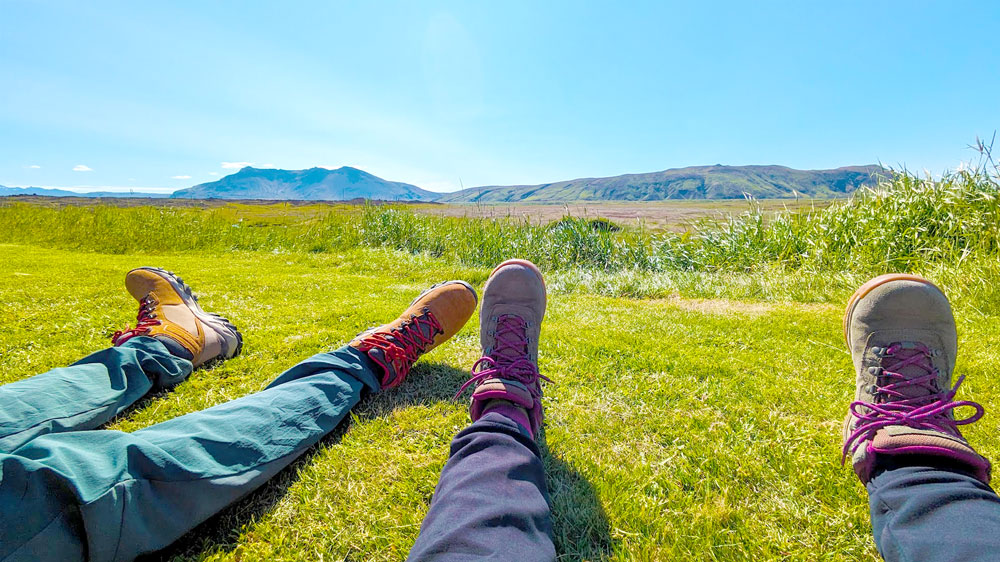 a couple relaxing on bright lush green grass somewhere in Iceland. You can only see the bottom of their legs, and their matching hiking shoes (in different colours).  They're on short grass, but taller unruly grass starts further down. In the distance are two mountain ranges - most likely volcanoes. They sky is pretty clear, with only a whisp of a white cloud. It's sunny and relaxing vibes. 