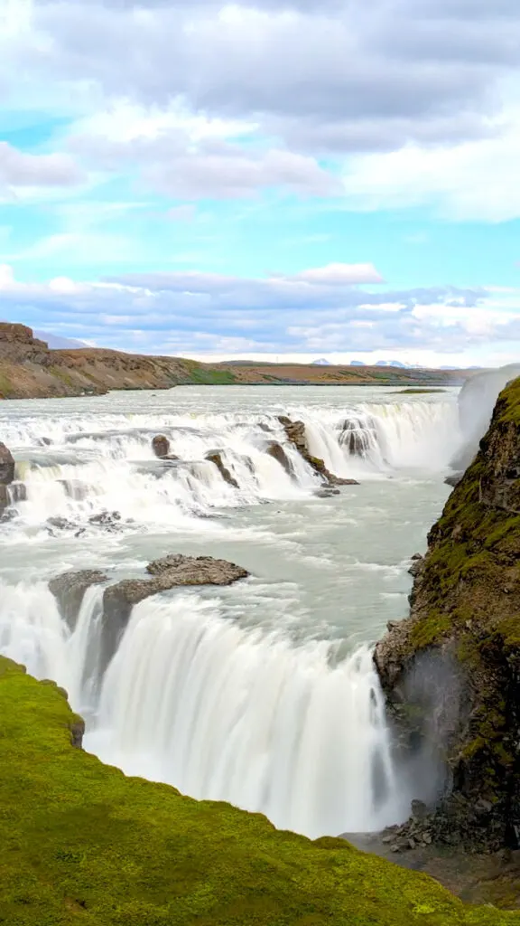 a vertical image showcasing a multilayered waterfall. The first drop is after a river flowing, it's not far, and the second drop is quickly after a sharp turn of the land. This is is longer and you can't see the bottom. It's a long exposure image, so the water looks very smooth and flowey. Green grass and rocks surround the falls. There are taller mountains are in the distance. The sky is broken up into thirds, a layer of clouds are nearest the horizon, then crisp blue sky, before another layer of clouds take over the top of the image. 