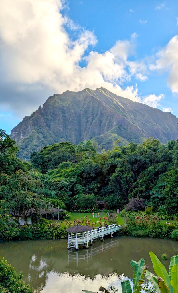 a relaxing jaw dropping view of the haiku gardens from above. There's a green pond, with the reflection of a white gazebo, which sits right above it going out into the middle pf the pond. Around it is green grass and lush green trees behind that. But the view is all about the taller than life mountain behind leading way up into the blue and cloudy sky. 