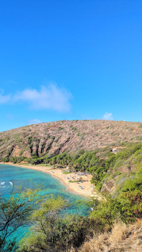 a view from above of the hanauma bay nature reserve. Its a beach with clear blue teal waters thats secluded between a rounded mountains around it. On the beach, you can see beach chairs and umbrellas already set up, and its really not busy, considering how popular it is